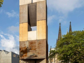 MAALOT Ein beeindruckendes Kunstobjekt aus rostigem Stahl erhebt sich vor der Kulisse des Kölner Doms. Der Turm wirkt monumental und kontrastiert mit dem blauen Himmel und den gotischen Kirchenspitzen.An impressive art object made of rusty steel rises up against the backdrop of Cologne Cathedral. The tower looks monumental and contrasts with the blue sky and the Gothic church spires.
