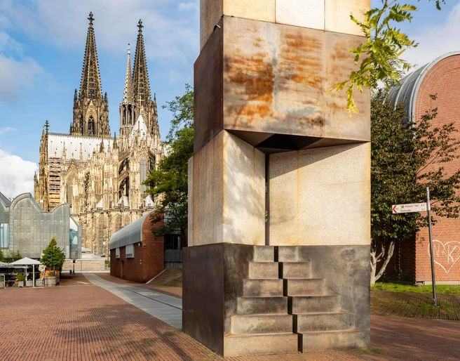 MAALOT Skulptur „MA'ALOT“ von Dani Karavan vor dem Kölner Dom bei sonnigem Himmel.Sculpture "MA'ALOT" by Dani Karavan in front of Cologne Cathedral under a sunny sky.