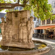 Ostermann Fountain Das Bild zeigt den Ostermann-Brunnen in Köln an einem sonnigen Tag. Mehrere Statuen umringen den rechteckigen Brunnen, aus dem Wasser sprudelt. Im Hintergrund sind ein Straßencafé und mehrere Gebäude mit Balkonen zu sehen, die von Bäumen beschattet werden.The picture shows the Ostermann fountain in Cologne on a sunny day. Several statues surround the rectangular fountain from which water gushes. A street café and several buildings with balconies shaded by trees can be seen in the background.