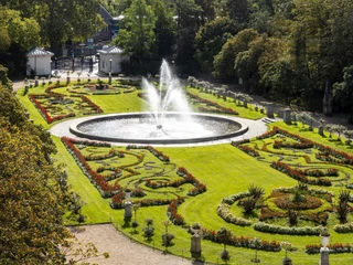 Flora and botanical garden Eine üppige Gartenanlage in symmetrischem Design zeigt einen zentralen Brunnen, umgeben von kunstvoll gestalteten Blumenbeeten, eingebettet in sattes Grün. Der Garten ist von dichtem Baumbestand umrahmt. A lush garden with a symmetrical design features a central fountain surrounded by artistically designed flowerbeds and lush greenery. The garden is surrounded by dense trees.