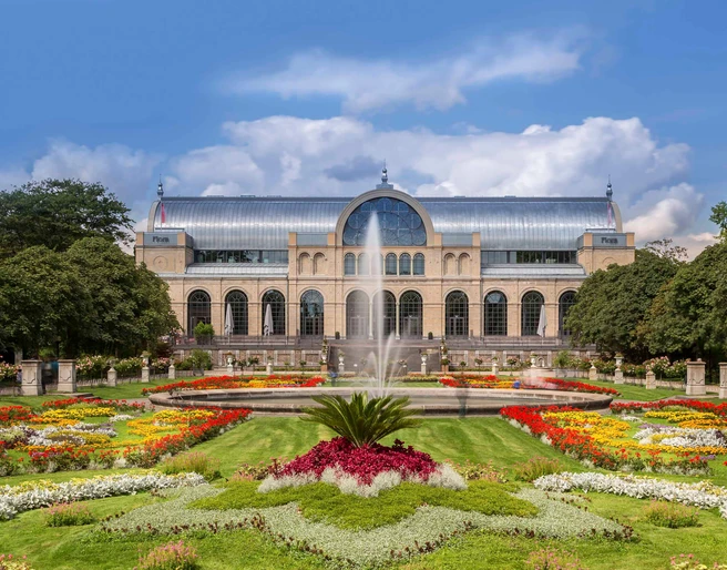 Flora and botanical garden Das Bild zeigt die historische Flora Köln, eine beeindruckende Orangerie mit gläserner Kuppel, eingebettet in farbenprächtige Blumenbeete und einen zentralen Springbrunnen.The picture shows the historic Flora Köln, an impressive orangery with a glass dome, embedded in colorful flowerbeds and a central fountain.