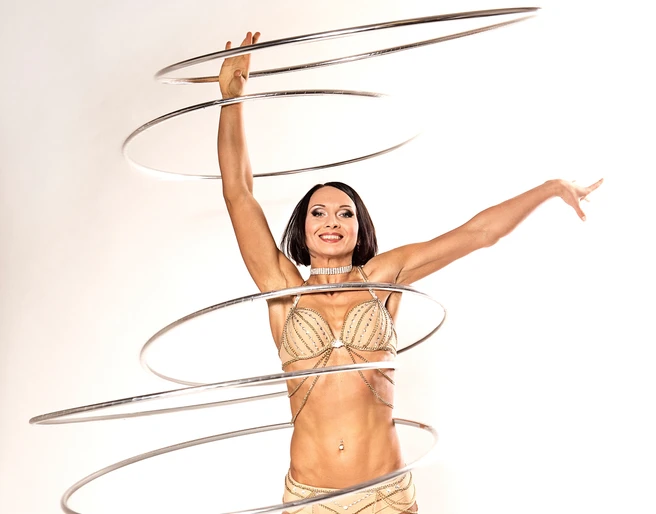 Olena 1.jpg Eine Frau in goldenen Outfits jongliert elegant acht Hula-Hoop-Reifen in einem Studio.