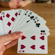 Canasta Eine Hand hält Karten mit Herzen, Karo, Pik und Kreuz. Zwei Personen spielen im Hintergrund.