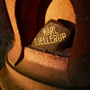 Geführte Wanderung zum Nobelpreisträger  Ein schwarzer Stein mit der goldenen Inschrift "Karl Gjellerup" liegt in einer rötlich beleuchteten Nische aus Stein.