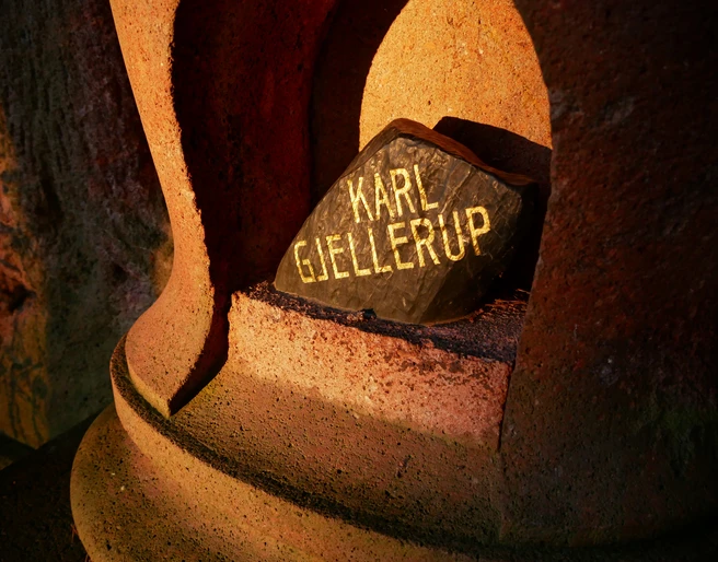 Geführte Wanderung zum Nobelpreisträger  Ein schwarzer Stein mit der goldenen Inschrift "Karl Gjellerup" liegt in einer rötlich beleuchteten Nische aus Stein.