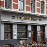 Ein Eingang des Lapidarium in Köln mit roten Ziegeln und blauen Fensterrahmen.Facade of a historic pub named "Lapidarium" with outdoor seating and brick architecture.