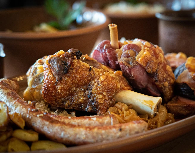 Haxenhaus Knusprige Schweinshaxe und Bratwurst auf einem rustikalen Holzteller mit Sauerkraut serviert.A rustic platter with a crispy pork knuckle, sausages, potatoes, and cabbage on a wooden table.