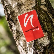Wegemarkierung vom neanderland STEIG Rote Wegemarkierung mit weißem Symbol und Schriftzug "neanderland STEIG" an einem Baumstamm.