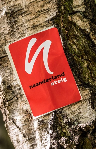 Wegemarkierung vom neanderland STEIG Rote Wegemarkierung mit weißem Symbol und Schriftzug "neanderland STEIG" an einem Baumstamm.
