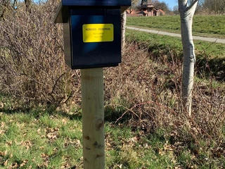 Stempelbox Foto Antje Geisler.jpg Das Bild zeigt eine blaue Stempelbox auf einem Holzpfosten in einer ländlichen Umgebung mit Bäumen und einer Windmühle im Hintergrund.
