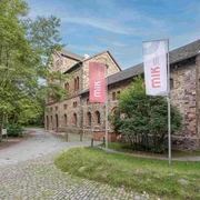 MIK Haseschachtgebäude 2022 Foto Frieso Gentsch_klein.jpg Historisches Fachwerkgebäude mit gemauerten Bruchsteinwänden, umgeben von Bäumen und zwei Fahnen.