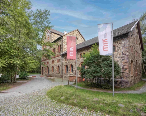 MIK Haseschachtgebäude 2022 Foto Frieso Gentsch_klein.jpg Historisches Fachwerkgebäude mit gemauerten Bruchsteinwänden, umgeben von Bäumen und zwei Fahnen.