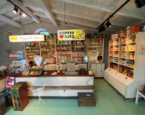 Tante_Emma_Laden_Klein.JPG Ein nostalgischer Tante-Emma-Laden mit Regalen voller bunter Produkte und einer einladenden Theke.