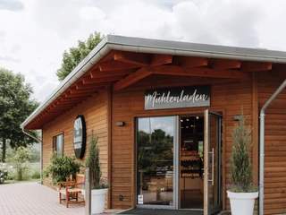 Ölmühle Solling Mülhenladen©Ölmühle Solling_Maike Wittreck.jpg Holzgebäude mit Mühlenladen-Schild, offenem Eingang und Pflanzen, umgeben von Bäumen und Wolkenhimmel.