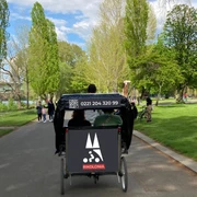 Rikolonia Picknick Rikscha Köln III.png Fahrradrikscha mit Fahrgästen in grünem Park, umgeben von Bäumen und Spaziergängern.Cycle rickshaw with passengers in a green park, surrounded by trees and walkers.