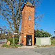Trafoturm Brokeloh Roter Backsteinturm mit bogenförmigen Fenstern, umgeben von einem Stromkasten und einer Sitzbank an der Straße.
