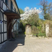 Gut Ehlenbeck in Haan Fachwerkhaus mit blühendem Baum vor der Tür, gepflasterter Weg und Gartenstühle unter blauem Himmel.
