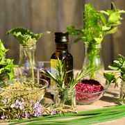 Chilli-Manufaktur-Lueneburger-Heide-Mitbringsel-herstellen.jpg Eine natürliche Kräuter- und Gewürzkomposition auf einem Holztisch im Freien. Verschiedene frische Kräuter wie Minze, Rosmarin und Schnittlauch stehen in kleinen Gläsern, daneben befinden sich Schalen mit getrockneten Kräutern und Gewürzen sowie eine Flasche Öl. Im Hintergrund ist ein rustikaler Holzzaun sichtbar. Die Szene strahlt eine natürliche, handgemachte Atmosphäre aus.