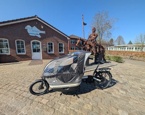 Lastenrad_Touristik Ein Lastenrad, platziert auf einem gepflasterten Hof vor der Apen Touristik, daneben eine Skulptur einer historischen Figur.
