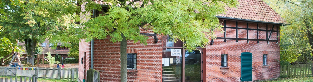 kuesterscheune_schmal.jpg Das Bild zeigt ein traditionelles Backsteingebäude, die Küsterscheune, umgeben von Bäumen im Garten.