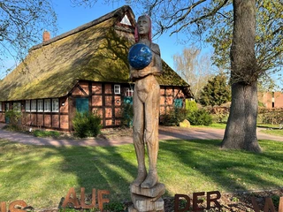 Weltenträgerin  Eine hölzerne Skulptur eines Menschen, der eine blaue Erdkugel hält, vor einem Fachwerkhaus.