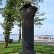 Skulptur aus dunklem Metall auf grünem Gras vor Bäumen und See. Im Hintergrund liegt ein Bootssteg.