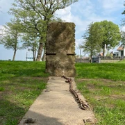 Steinhude Skulpturenpromenade: Aufrecht stehende Steinplatte mit Ketten auf einer Wiese am Seeufer.