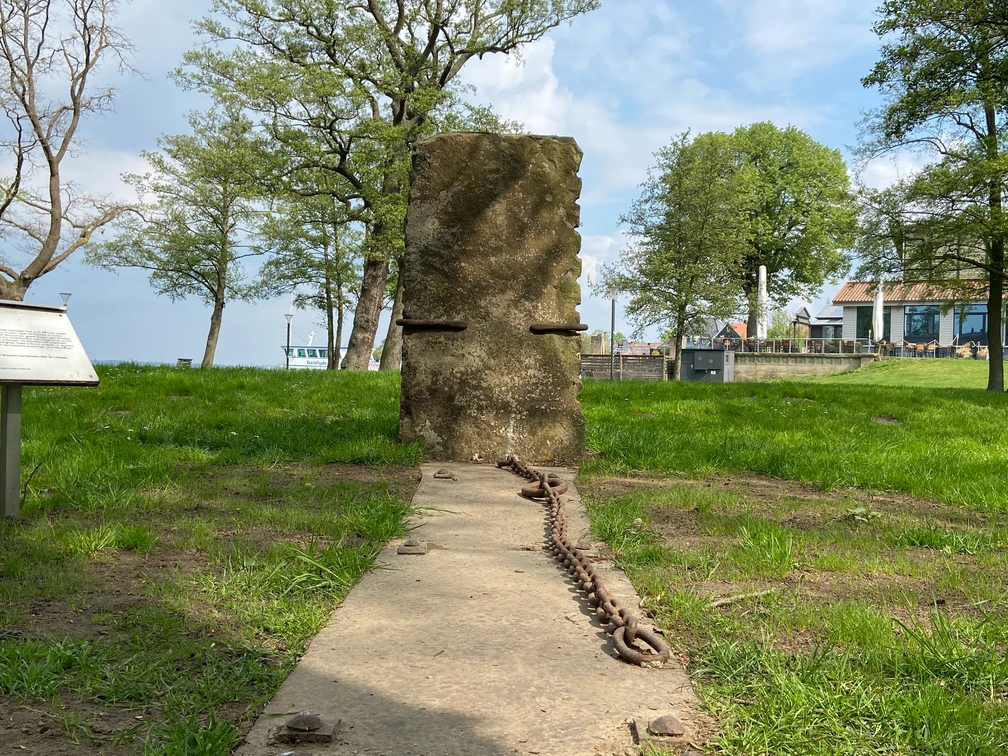 Hinführung-Skulptur-Skulpturenpromenade-Steinhude3 Steinhude Skulpturenpromenade: Aufrecht stehende Steinplatte mit Ketten auf einer Wiese am Seeufer.