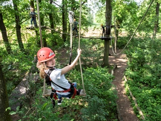 Kletterpark-Mardorf_dsc_7255_klein.jpg Eine junge Frau schwingt sich in einem Kletterpark zwischen Bäumen, gesichert mit Helm und Gurt.