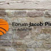 forum-jacobpins-schild Schild aus Metall mit orangefarbenem Symbol und schwarzer Schrift auf aus Stein gemauerter Wand.