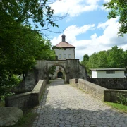 Historisches Gut mit Brücke und Fachwerkgebäude umgeben von üppigem Grün unter blauem Himmel.