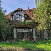 Fachwerkhaus mit rotem Ziegeldach, umgeben von grünen Bäumen, mit einem rustikalen Holzschild davor.