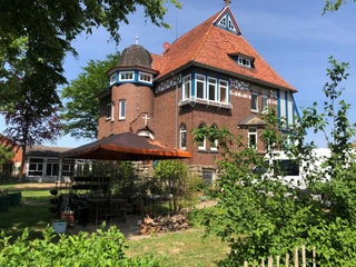 Pilgerherberge Altes Pfarrhaus Backsteinbau mit rotem Ziegeldach, weißen Gauben und Erkern in grüner Gartenumgebung.
