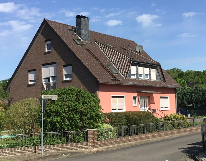 FeWo Süchting Ein zweistöckiges Haus mit roter Fassade und braunen Dächern, umgeben von grünen Büschen und Bäumen.