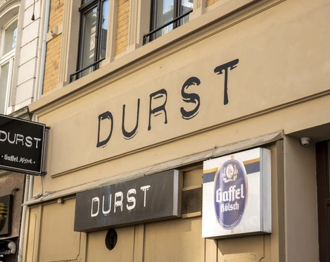 Durst Leuchtreklame „Durst“ auf Gebäudefront mit Gaffel Kölsch-Schild an einer historischen Straße.Illuminated sign reading ‘Durst’ (Thirst) on the front of a building with a Kölsch sign on a historic street.