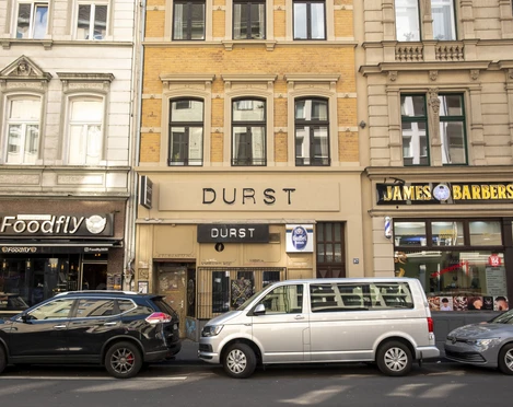 Durst Die Straßenszene in Köln zeigt das "Durst"-Gebäude zwischen einem Friseursalon und einem Restaurant, mit geparkten Autos entlang der Straße. Der Schriftzug "DURST" prägt die Fassade in klarer, moderner Schrift. The street scene in Cologne shows the ‘Durst’ building between a hairdressing salon and a restaurant, with parked cars along the street. The word ‘DURST’ dominates the façade in clear, modern lettering.