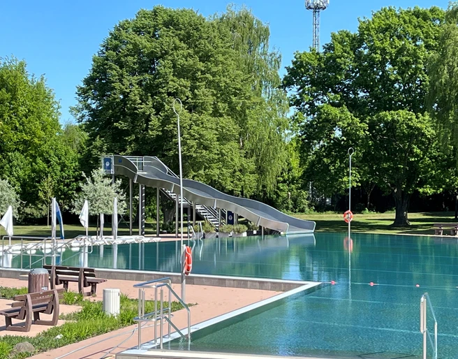 Freibad Achim Freibad mit großer Wasserrutsche im Grünen, umgeben von Bäumen und klarblauem Himmel.