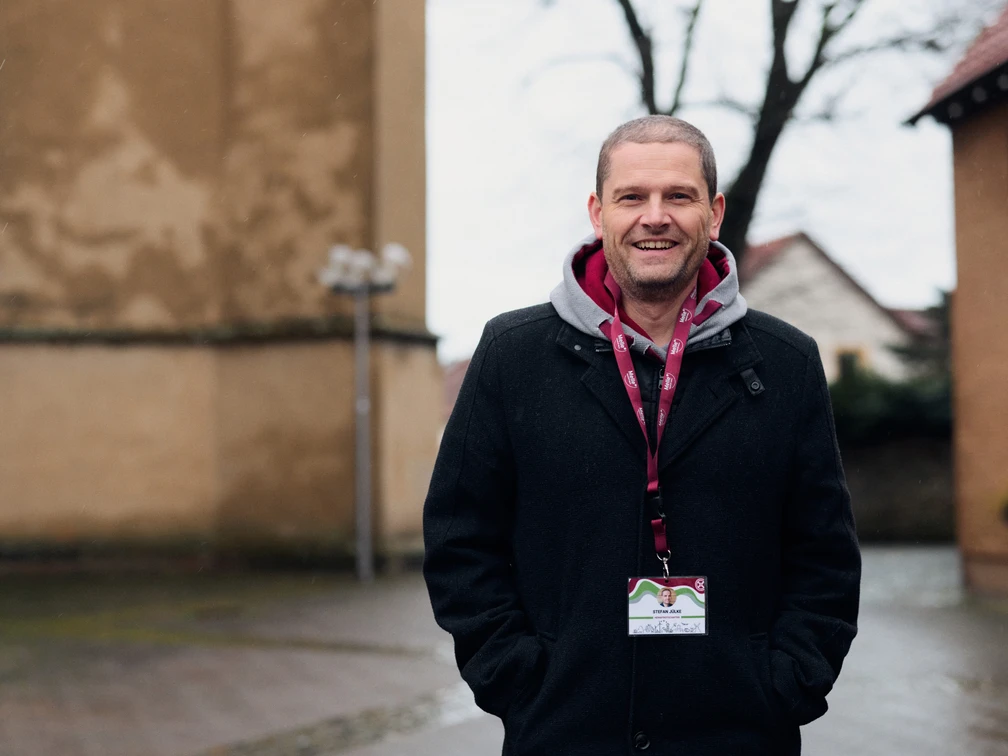 Stadtführer Stefan Jülke.jpg Ein lächelnder Mann steht in einem Innenhof vor einer historischen Gebäudefassade. Er trägt einen schwarzen Mantel und hat eine rote Umhängeleine um den Hals. Die Umgebung wirkt ruhig und fachwerkartig, mit vereinzelt stehenden Laternen und kahlen Bäumen im Hintergrund. Ein einladender Tag für eine geführte Stadterkundung.