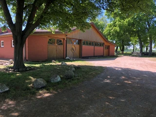 Reitstall zum Eichenhof (5).jpg Ein Reitstall mit rotem Ziegelgebäude und einer Holzfassade, umgeben von großen, Schatten spendenden Bäumen.