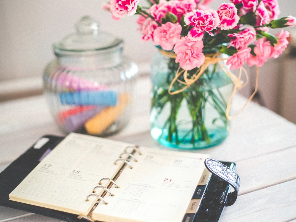 Bullet Journal Ein geöffneter Planer liegt auf einem Tisch neben einem Kerzenglas und einem Glas mit rosa Blumen.