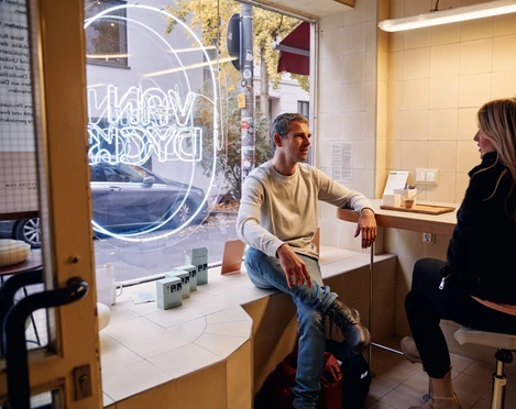 VAN DYCK Espressobar Ein Mann und eine Frau sitzen entspannt in der VAN DYCK Espressobar, hinter ihnen ist ein großes, weiß beleuchtetes Fenster mit dem Logo der Bar zu sehen. Die Atmosphäre wirkt warm und einladend.A man and a woman are sitting relaxed in the VAN DYCK espresso bar, behind them a large, brightly lit window displays the café's logo. The atmosphere appears warm and inviting.