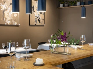 Pottkind Das Bild zeigt einen eleganten, modernen Restaurant-Innenraum mit stilvoller Tischdekoration, künstlerischen Wandbildern und einer harmonischen Beleuchtung. Im Vordergrund steht ein dekorativer Blumenstrauß in einer Glasvase auf einem hölzernen Bartresen.The image shows an elegant, modern restaurant interior featuring tasteful table decor, artistic wall prints, and harmonious lighting. In the foreground, a decorative flower arrangement in a glass vase is placed on a wooden bar counter.