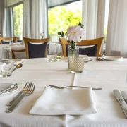 Zur Tant Ein elegant gedeckter Tisch in einem hellen, lichtdurchfluteten Restaurant. Weiße Tischdecke mit Besteck, Gläsern und einem Blumenvasen-Arrangement.An elegantly set table in a bright, light-filled restaurant. White tablecloth with cutlery, glasses, and a floral vase arrangement.