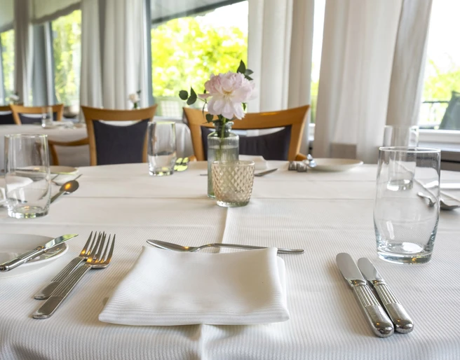 Zur Tant Ein elegant gedeckter Tisch in einem hellen, lichtdurchfluteten Restaurant. Weiße Tischdecke mit Besteck, Gläsern und einem Blumenvasen-Arrangement.An elegantly set table in a bright, light-filled restaurant. White tablecloth with cutlery, glasses, and a floral vase arrangement.