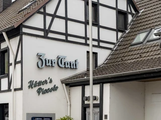 Zur Tant Ein Fachwerkhaus mit dem Schriftzug "Zur Tant". Das Gebäude ist weiß mit dunklen Holzbalken, einem Schornstein, und es gibt einen Eingang mit einer Tür und Schaufenstern.A half-timbered house with the inscription "Zur Tant". The building is white with dark timber beams, a chimney, and has an entrance with a door and shop windows.