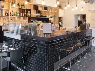 maiBeck Einladende Theke in einem modernen Restaurant. Die Theke ist mit glänzenden schwarzen Fliesen verkleidet und umrahmt eine Auswahl an Weinen, Gläsern und dekorativen Elementen. An der Theke befinden sich stilvolle Barhocker, während im Vordergrund ein gedeckter Tisch für zwei Gäste bereitsteht. Der Raum ist durch sanfte Beleuchtung und hängende Glühbirnen an der Decke atmosphärisch gestaltet.Welcoming counter in a modern restaurant. The counter is clad with shiny black tiles, framing a selection of wines, glasses, and decorative elements. Stylish bar stools are arranged at the counter, while in the foreground, a set table awaits two guests. The space is atmospherically designed with soft lighting and hanging bulbs suspended from the ceiling.