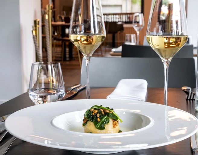 maiBeck Elegantes Restaurantinterieur mit stilvoll gedecktem Tisch, fokussiert auf ein exquisites Gericht mit frischen, grünen Zutaten, flankiert von zwei Gläsern Weißwein. Elegant restaurant interior featuring a stylishly set table, centered around an exquisite dish with fresh, green ingredients, accompanied by two glasses of white wine.