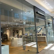 COMSPOT City Galerie Wolfsburg  Die Fassade eines modernen Elektronikgeschäfts mit Glasfront, benannt "COMSPOT", im Einkaufszentrum. Innen sieht man helle Holzfußböden und Ausstellungsflächen.