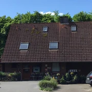 Ein rotes Backsteinhaus mit großem Satteldach und kleinen Oberlichtern vor üppig grüner Waldkulisse.