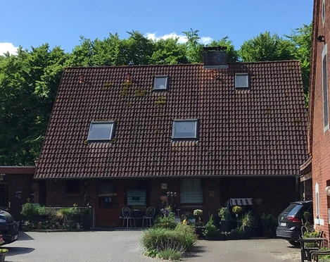 Zur Freilichtbühne Ein rotes Backsteinhaus mit großem Satteldach und kleinen Oberlichtern vor üppig grüner Waldkulisse.
